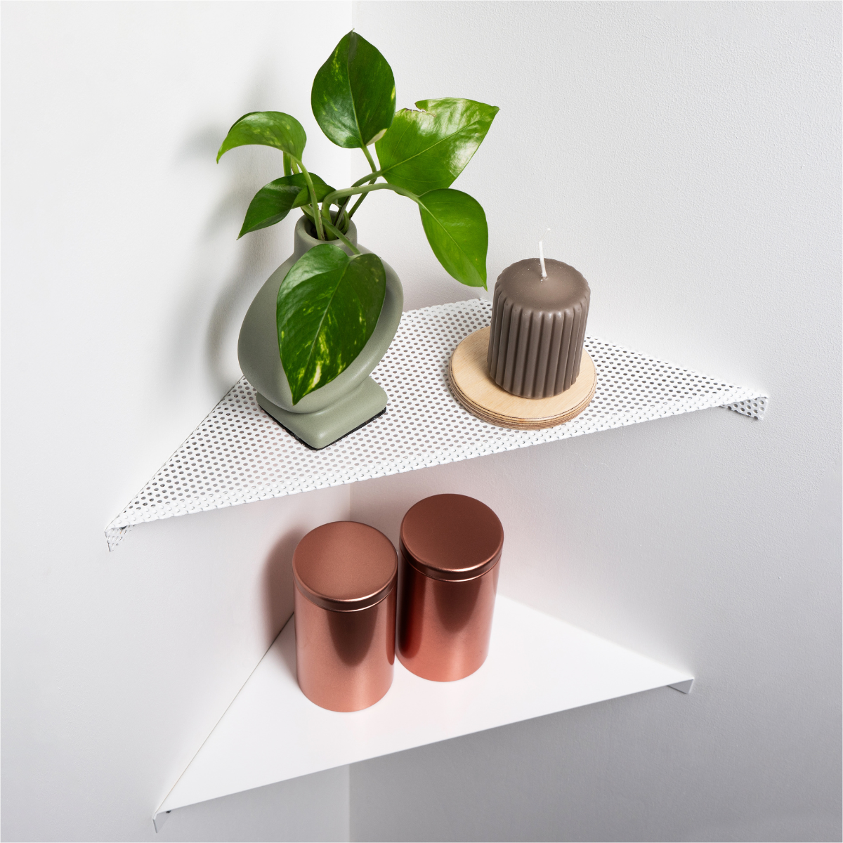 Two white corner shelves, one perforated, holding a green plant, brown candle on a wooden coaster, and two copper-colored containers.
